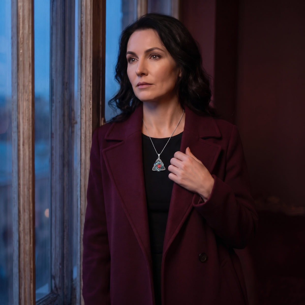 Woman in a wine coat wearing the witch knot by a window at dusk